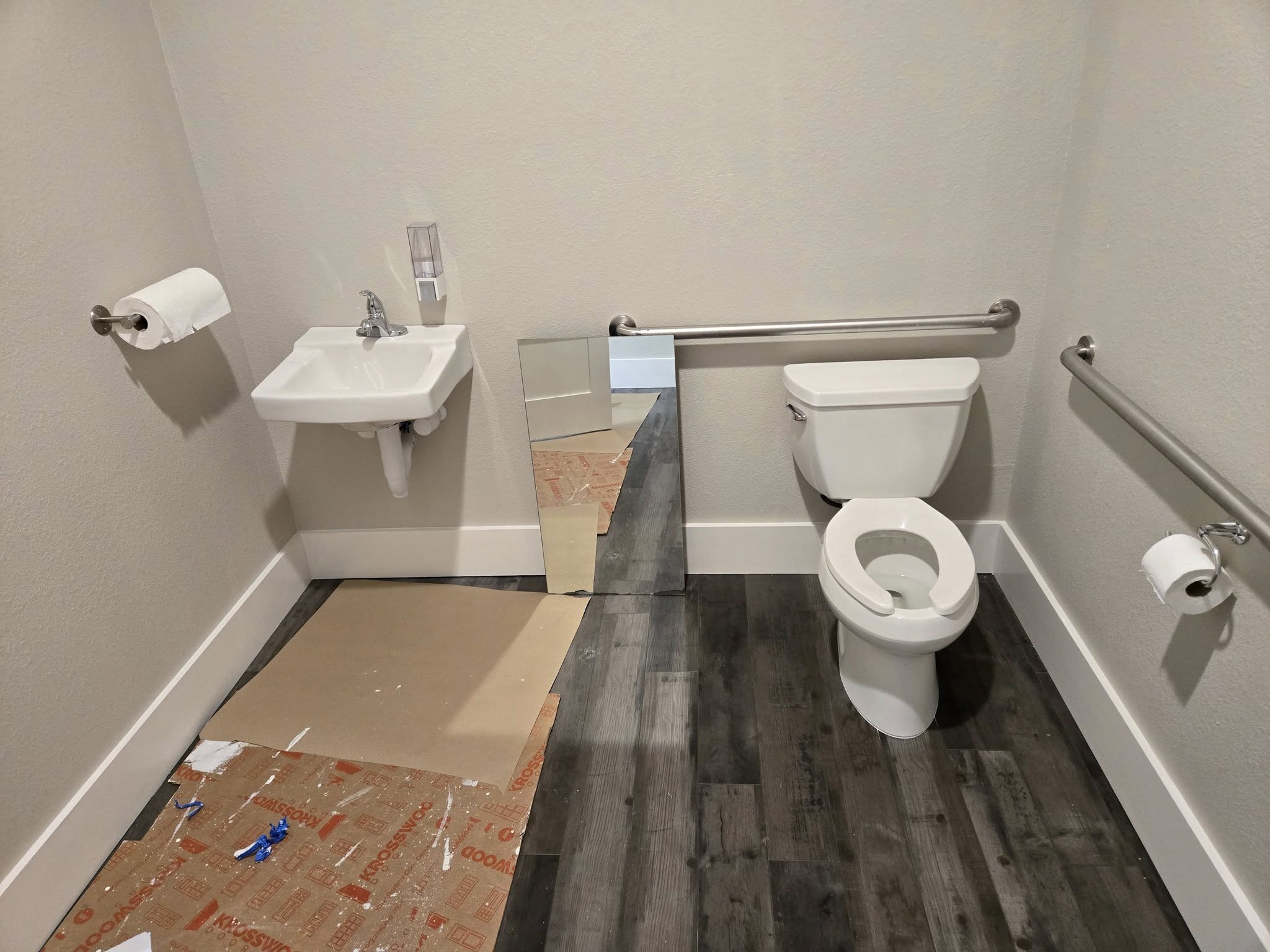 Commercial restroom plumbing in progress with toilet, sink, and mirror staged for install