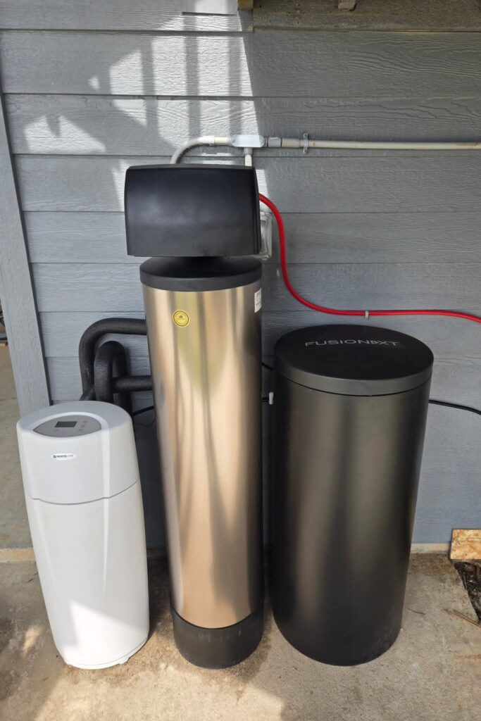 Outdoor water softener and filtration tanks installed beside a home with connected water lines.